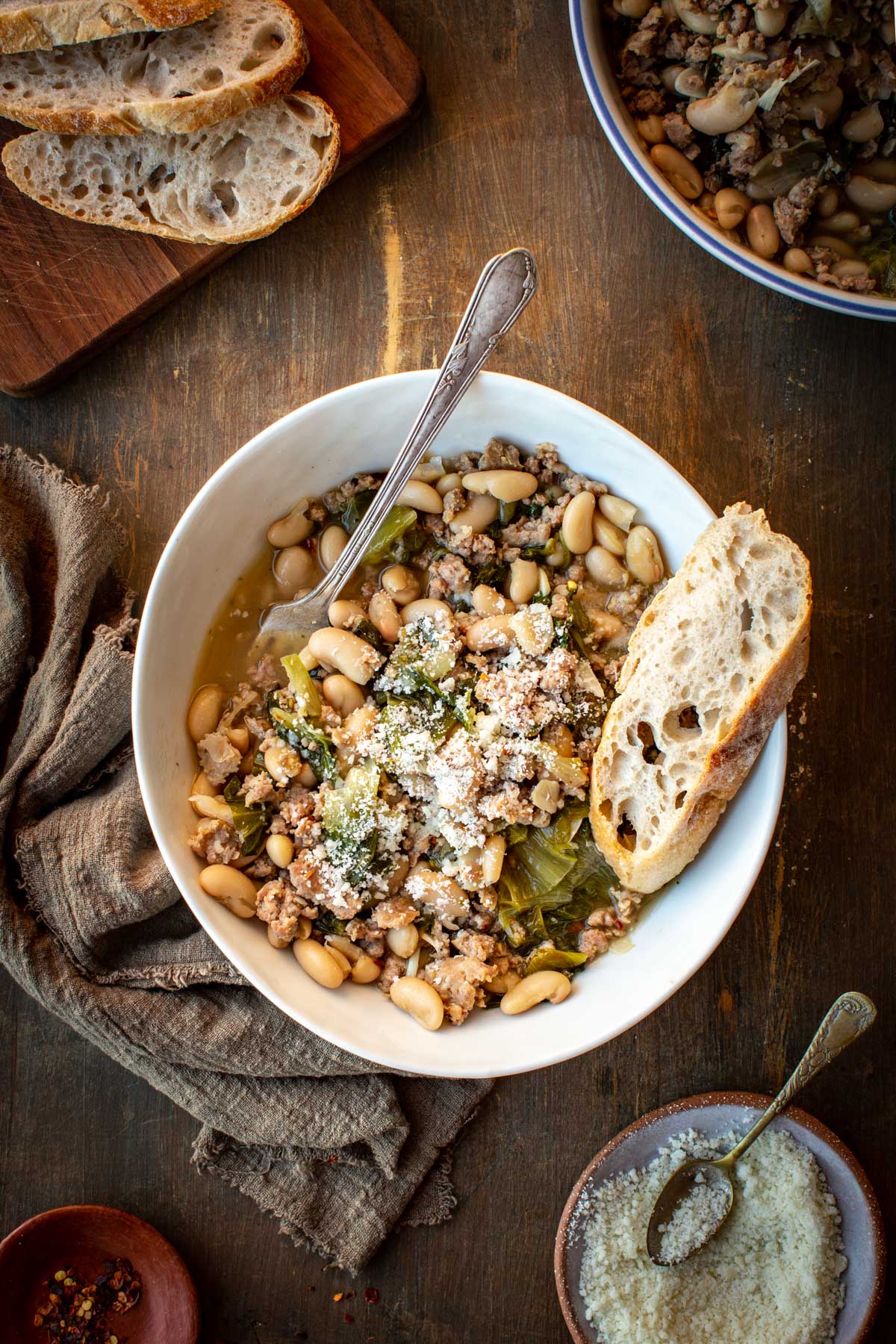 Hands holding a bowl of sausage, escarole, and white bean stew while tearing bread for dipping.