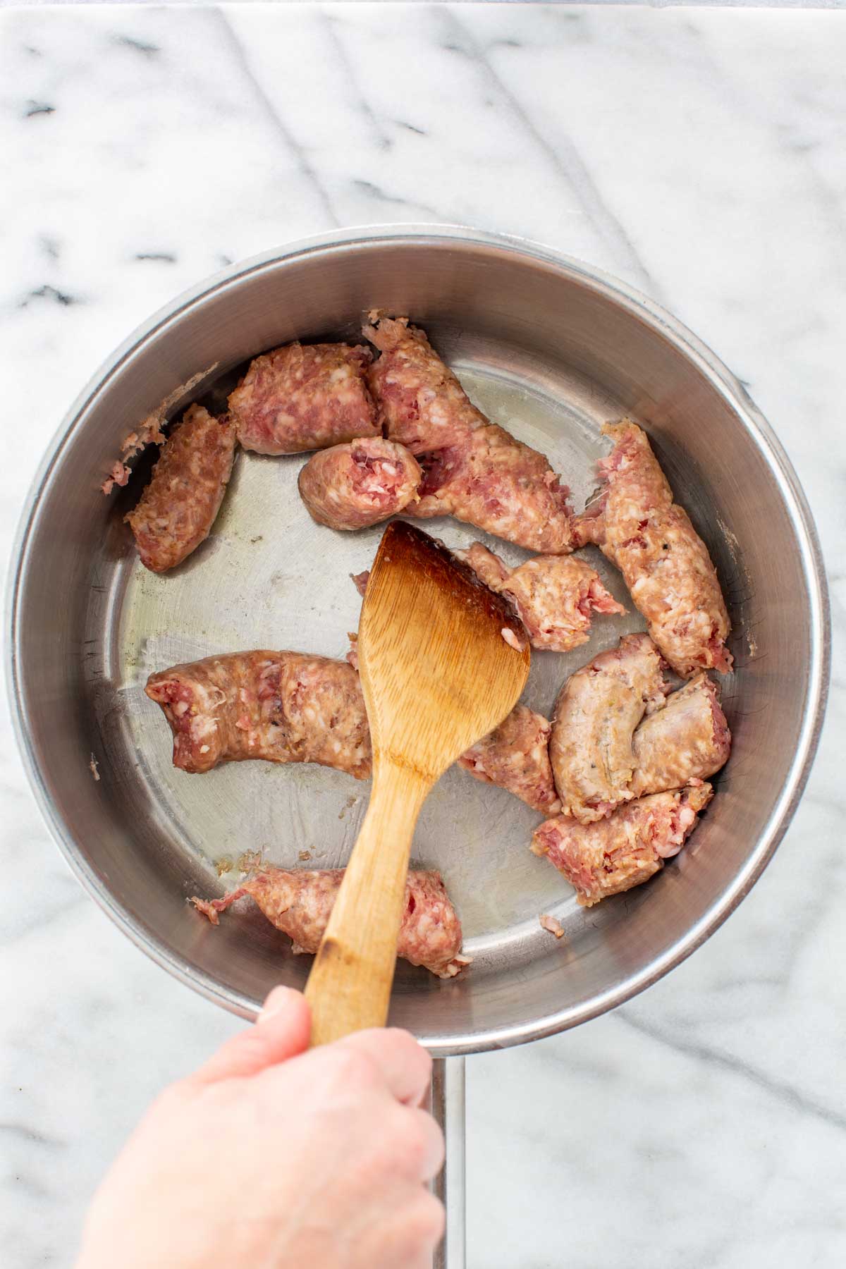 Italian sausage links broken into chunks and browned in a stainless steel pot with olive oil, stirred with a wooden spoon.
