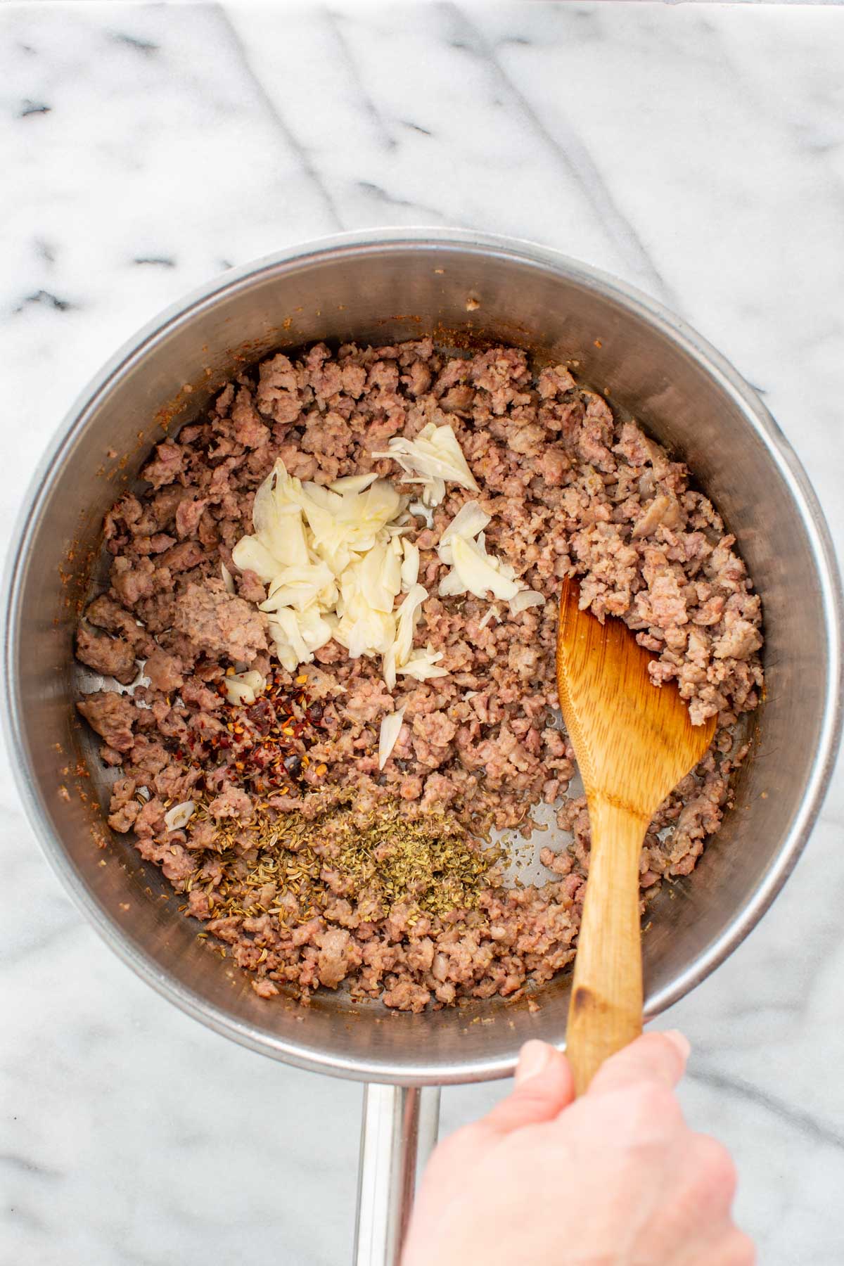 Browned sausage crumbles in the pot with sliced garlic, fennel seed, oregano, and red pepper flakes added on top before stirring.