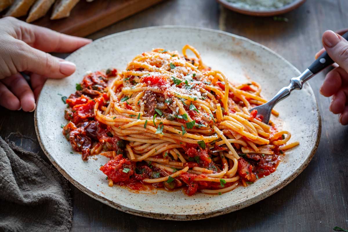 Close up of spaghetti puttanesca showing strands of pasta coated in tomato sauce with olives and capers.