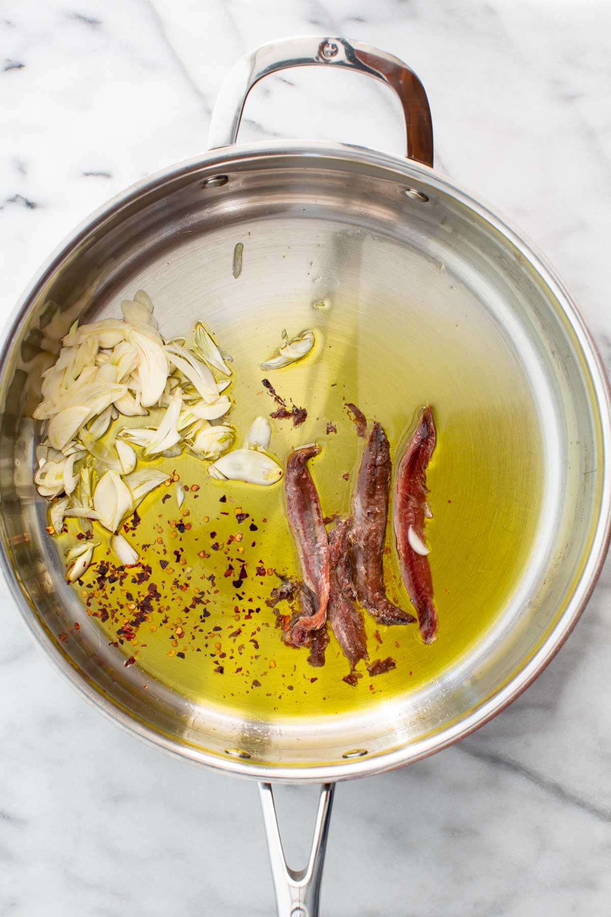 Olive oil heating in a stainless steel pan with sliced garlic, anchovy fillets, and red pepper flakes just beginning to melt and sizzle.