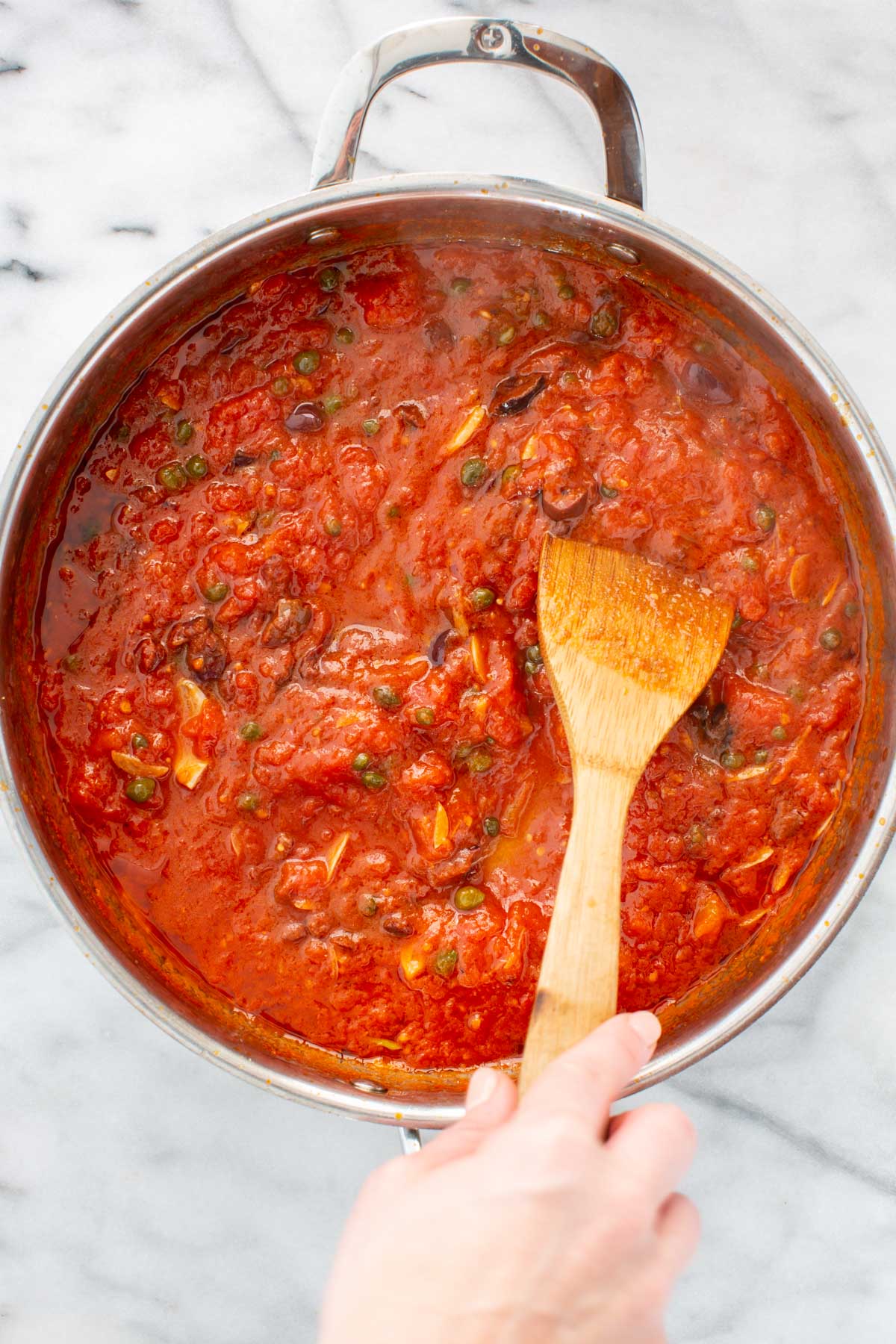 Tomato sauce simmering with olives, capers, and garlic, thickening slightly in the pan.