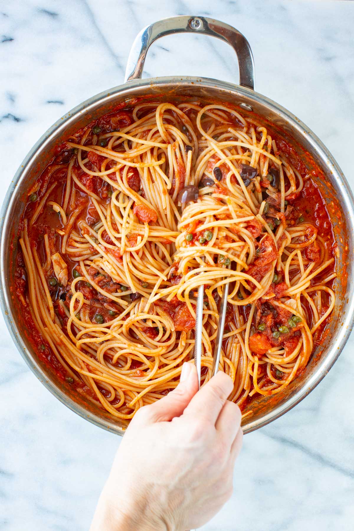 Cooked spaghetti tossed directly into the puttanesca sauce using tongs.
