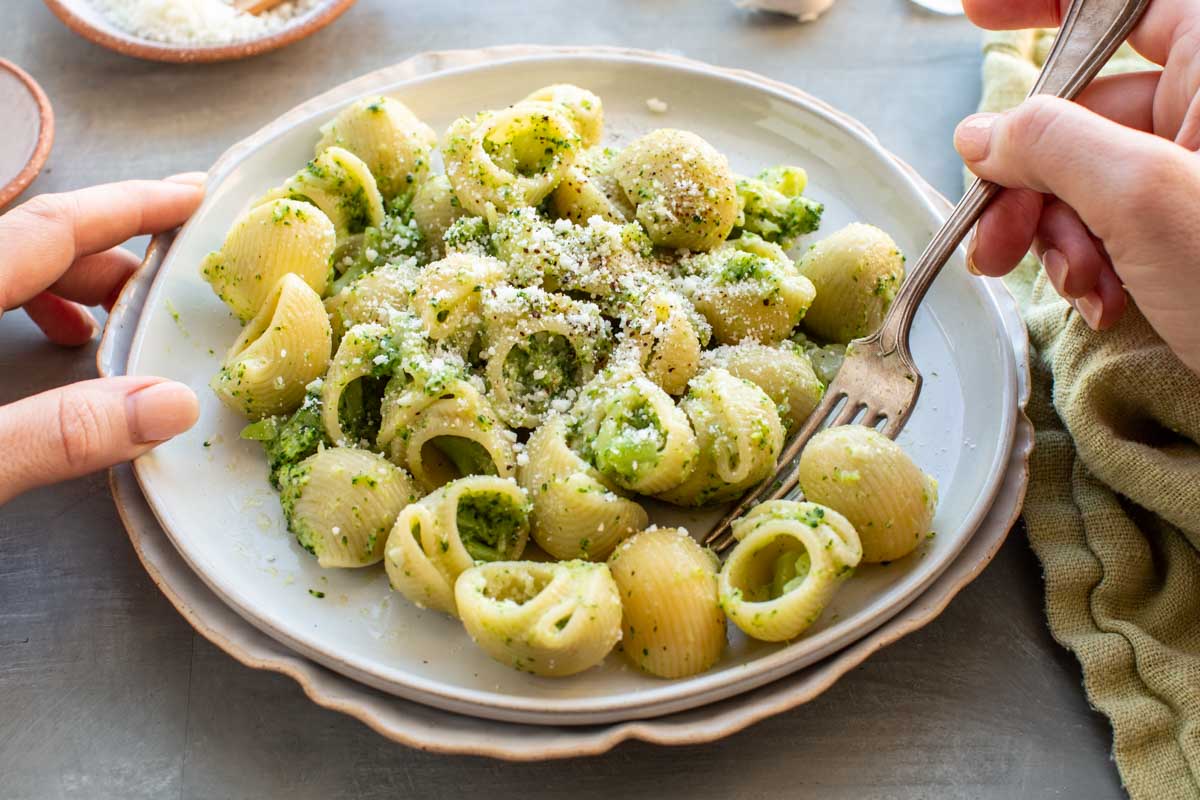 Side view of hands eating a plate of broccoli pasta with a fork.