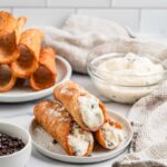Filled cannoli on a white plate with extra ricotta cream and mini chocolate chips in bowls behind them.