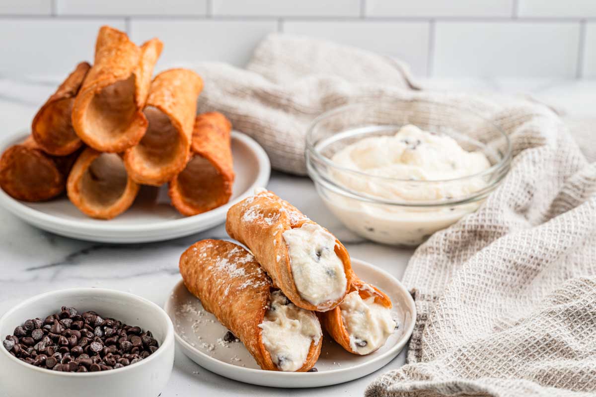 Filled cannoli on a white plate with extra ricotta cream and mini chocolate chips in bowls behind them.