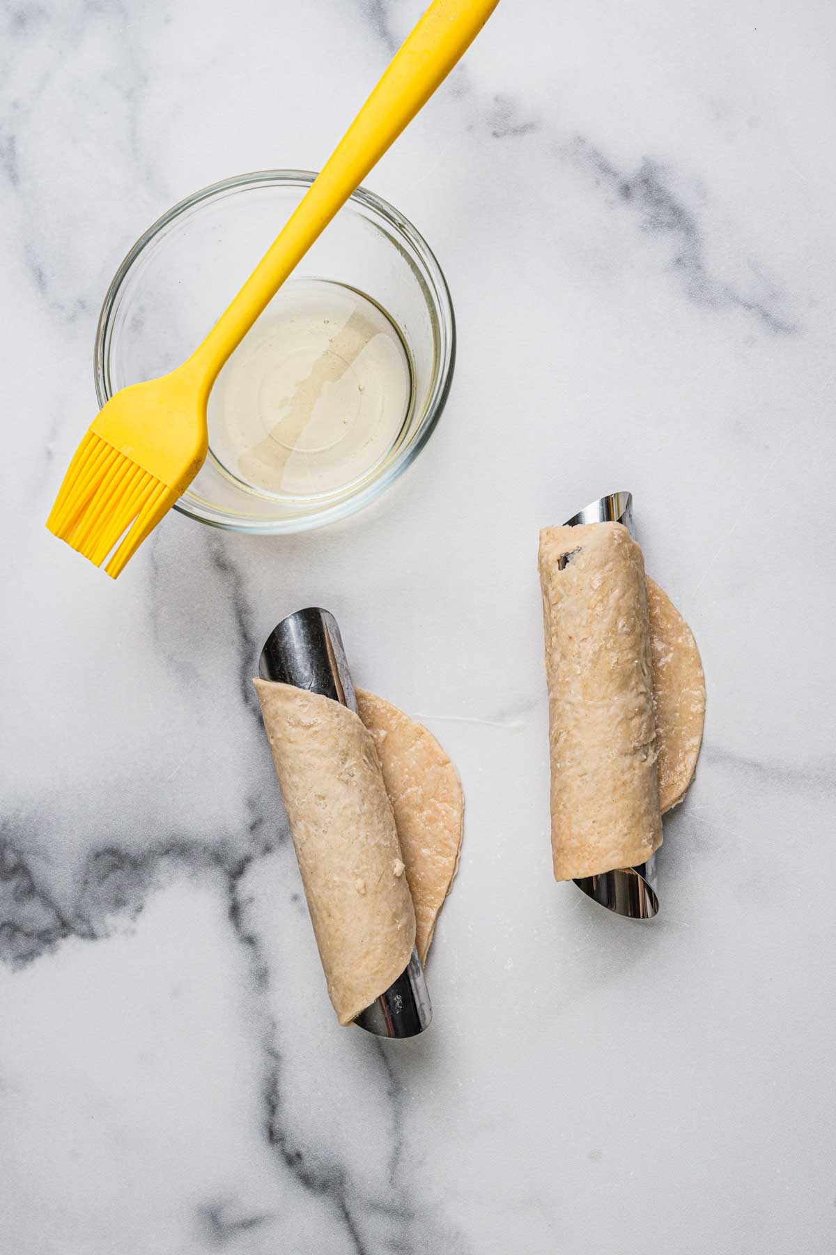 Unfried cannoli dough wrapped around stainless steel molds beside a pastry brush and bowl of egg wash on a marble countertop.