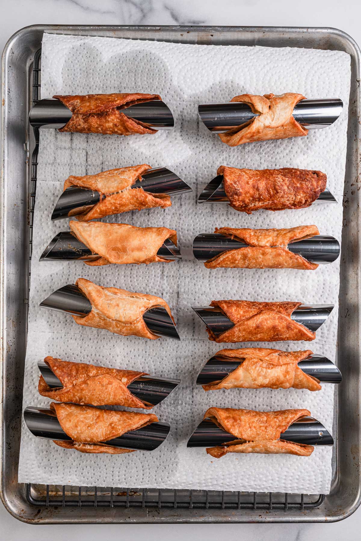 Golden brown cannoli shells cooling on stainless steel molds set over paper towels on a baking sheet.
