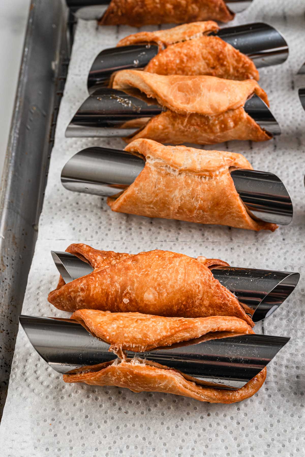 Crisp, blistered cannoli shells wrapped around stainless steel tubes, draining on a paper towel lined tray.