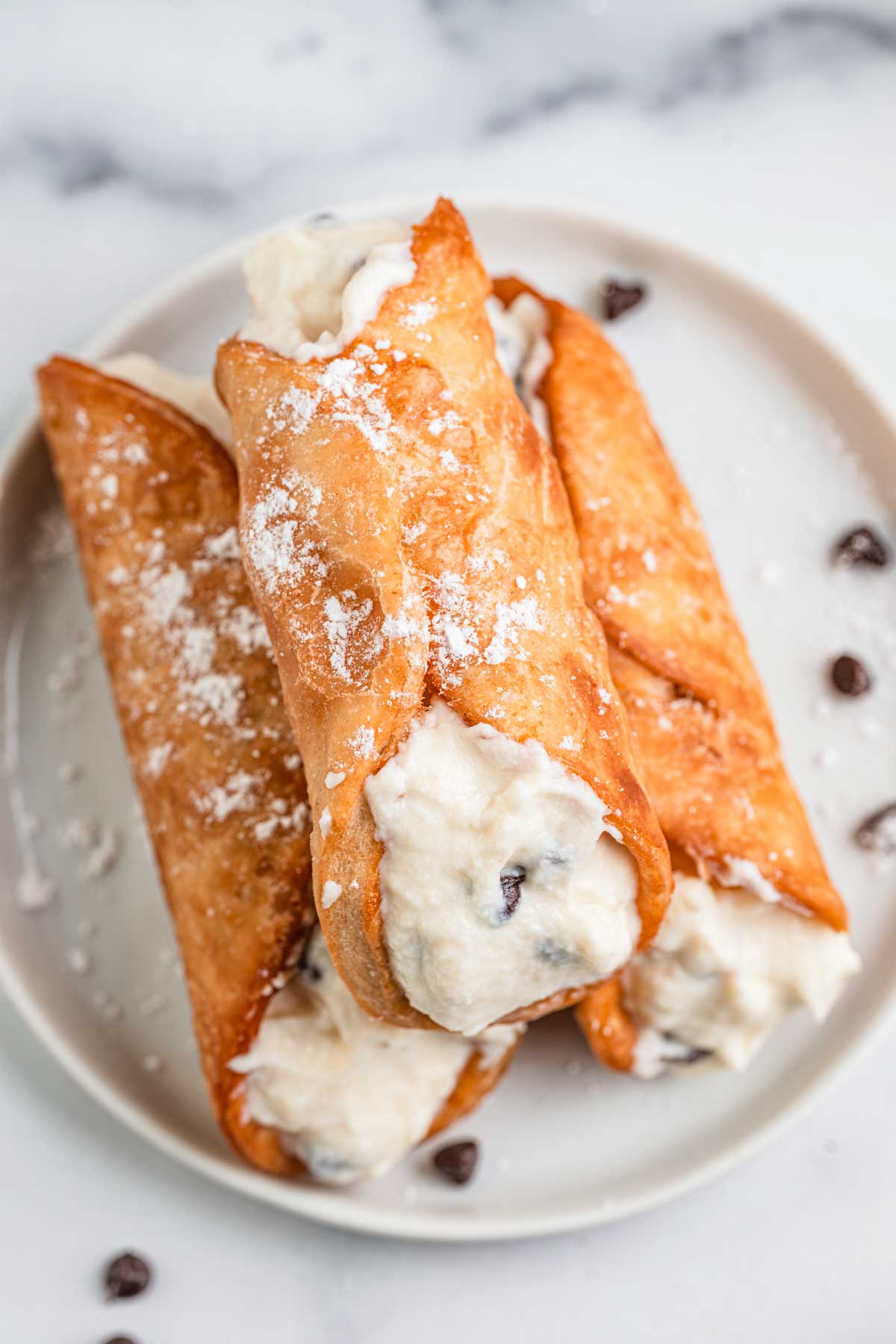 Three cannoli filled with sweet ricotta and mini chocolate chips, lightly dusted with powdered sugar on a plate.