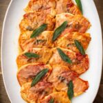 Overhead view of chicken saltimbocca on a white oval platter, topped with prosciutto and fresh sage leaves in a light buttery pan sauce.
