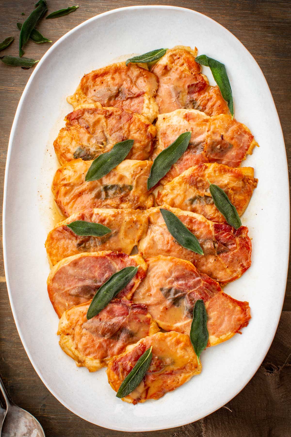 Overhead view of chicken saltimbocca on a white oval platter, topped with prosciutto and fresh sage leaves in a light buttery pan sauce.