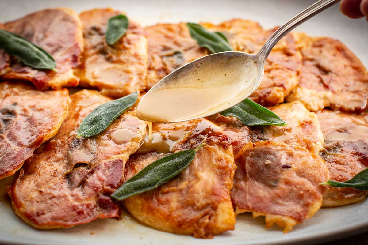 Close-up of chicken saltimbocca with prosciutto and sage as a spoon drizzles glossy white wine butter sauce over the top.