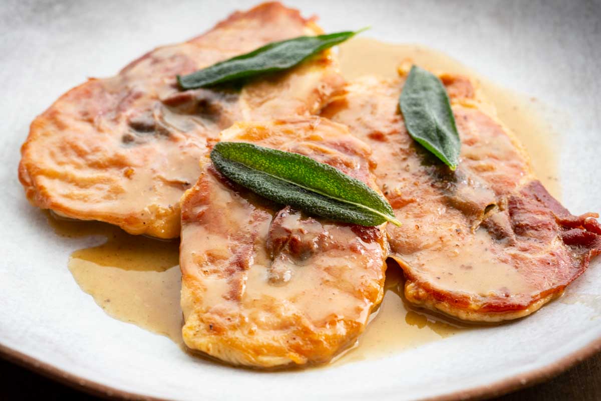 Three pieces of chicken saltimbocca with prosciutto and sage on a white plate, coated in a light buttery wine sauce.