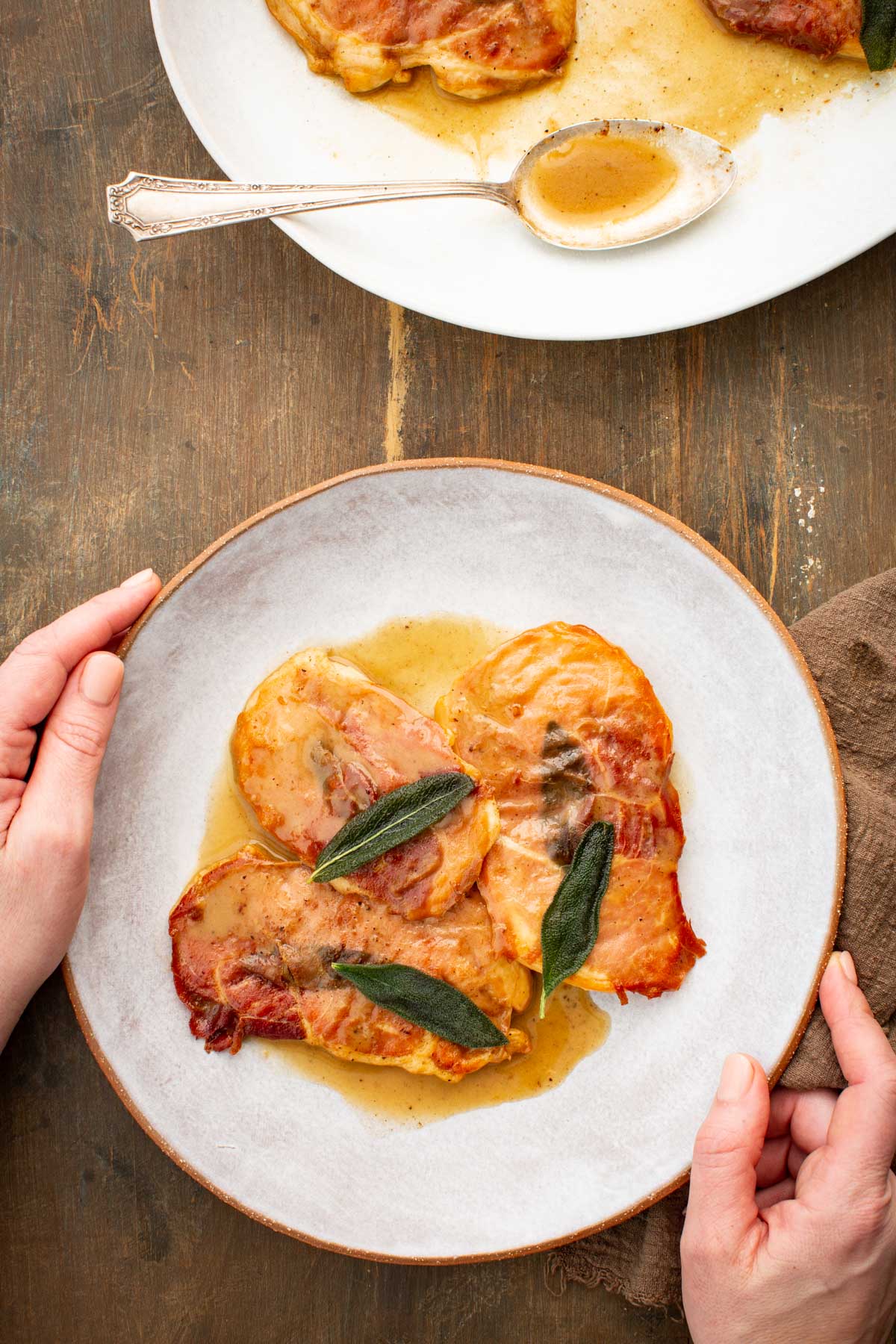 Plate of chicken saltimbocca topped with prosciutto and sage in a butter sauce, held over a rustic wooden table with serving platter in background.