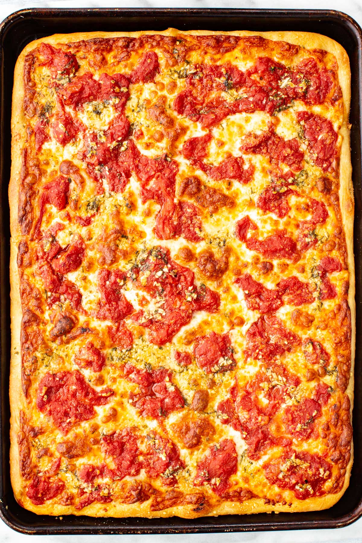 Overhead view of a rectangular grandma pizza baked in a sheet pan with a golden, crisp crust, melted cheese, chunky tomato sauce, and dried herbs scattered on top.