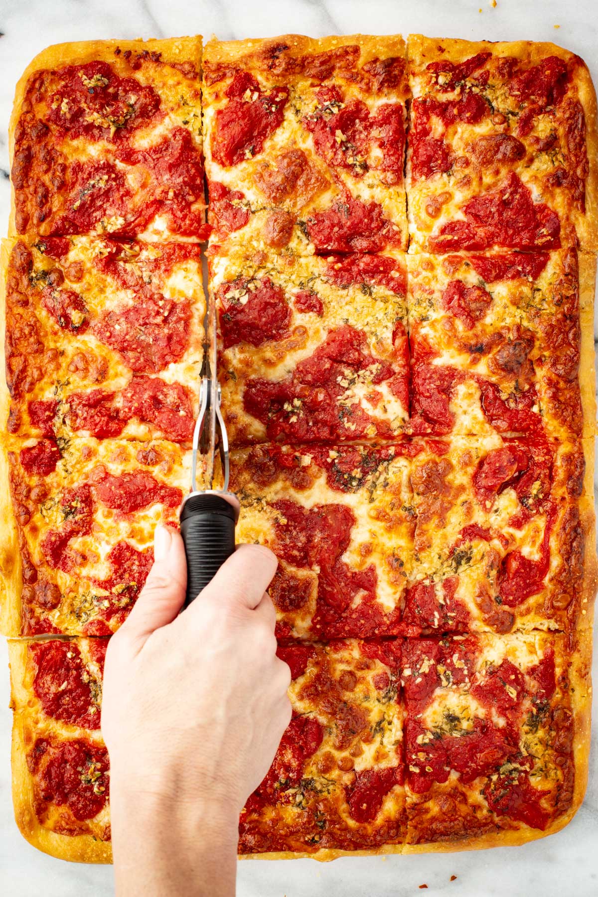 A hand using a pizza cutter to slice a grandma pizza into square pieces while it sits in the baking pan.