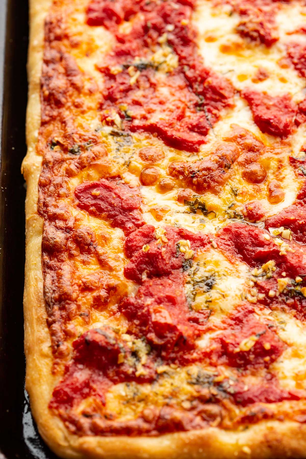 Close up of a grandma pizza showing the thick, golden crust, bubbly cheese, and pockets of chunky tomato sauce.