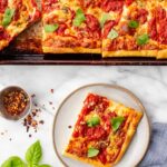 Overhead view of grandma pizza cut into squares on a well-worn sheet pan, topped with fresh basil leaves.