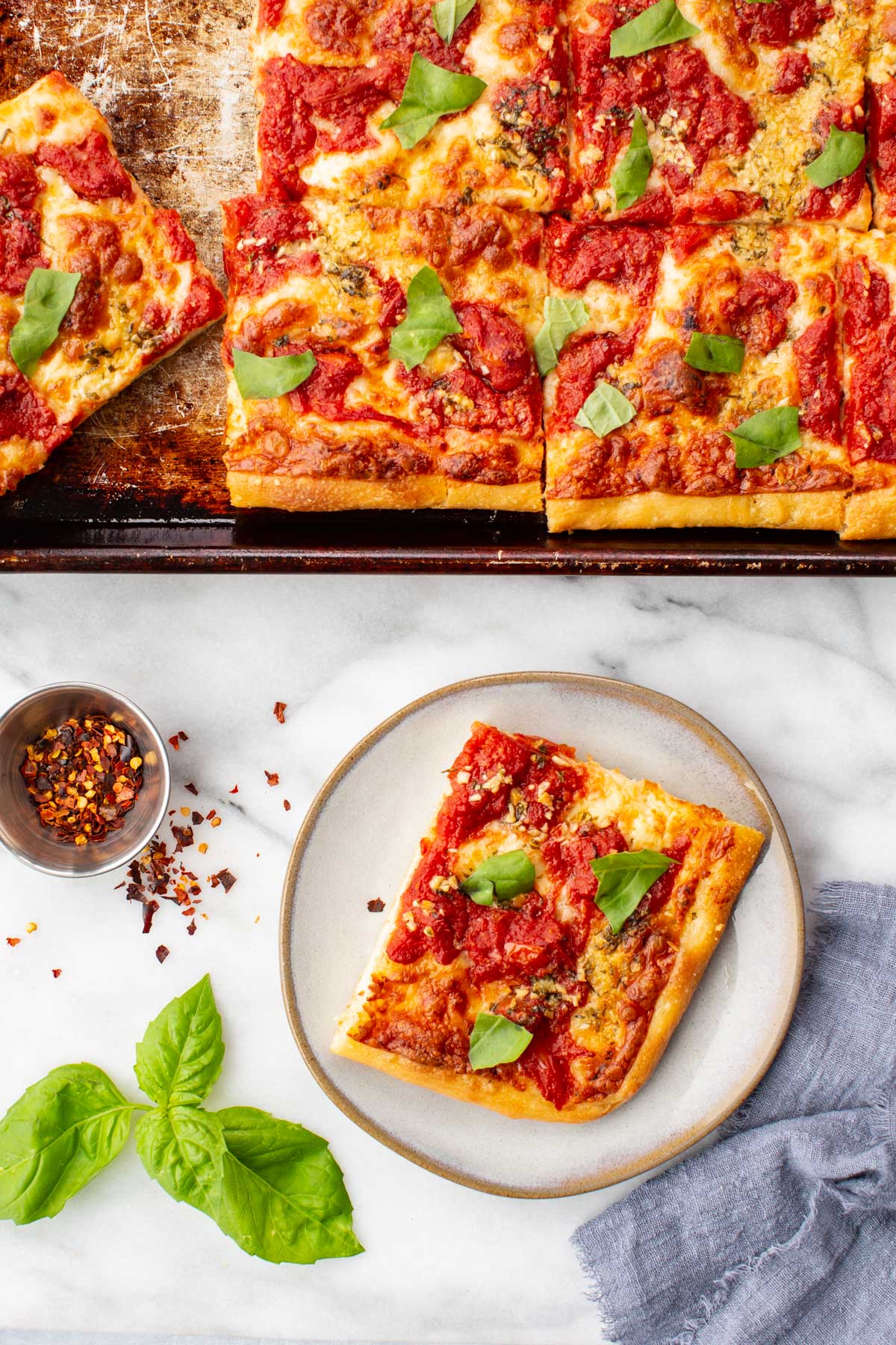 Overhead view of grandma pizza cut into squares on a well-worn sheet pan, topped with fresh basil leaves.