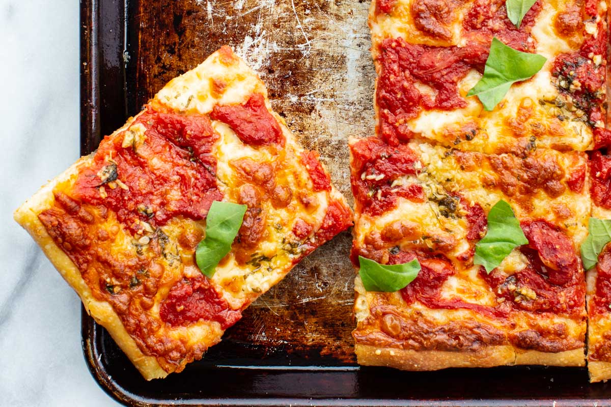 A single square slice of grandma pizza served on a plate, garnished with fresh basil, with chili flakes and herbs nearby.