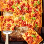 Multiple square slices of grandma pizza arranged on a baking tray and plate, ready to serve, with fresh basil and red pepper flakes alongside.