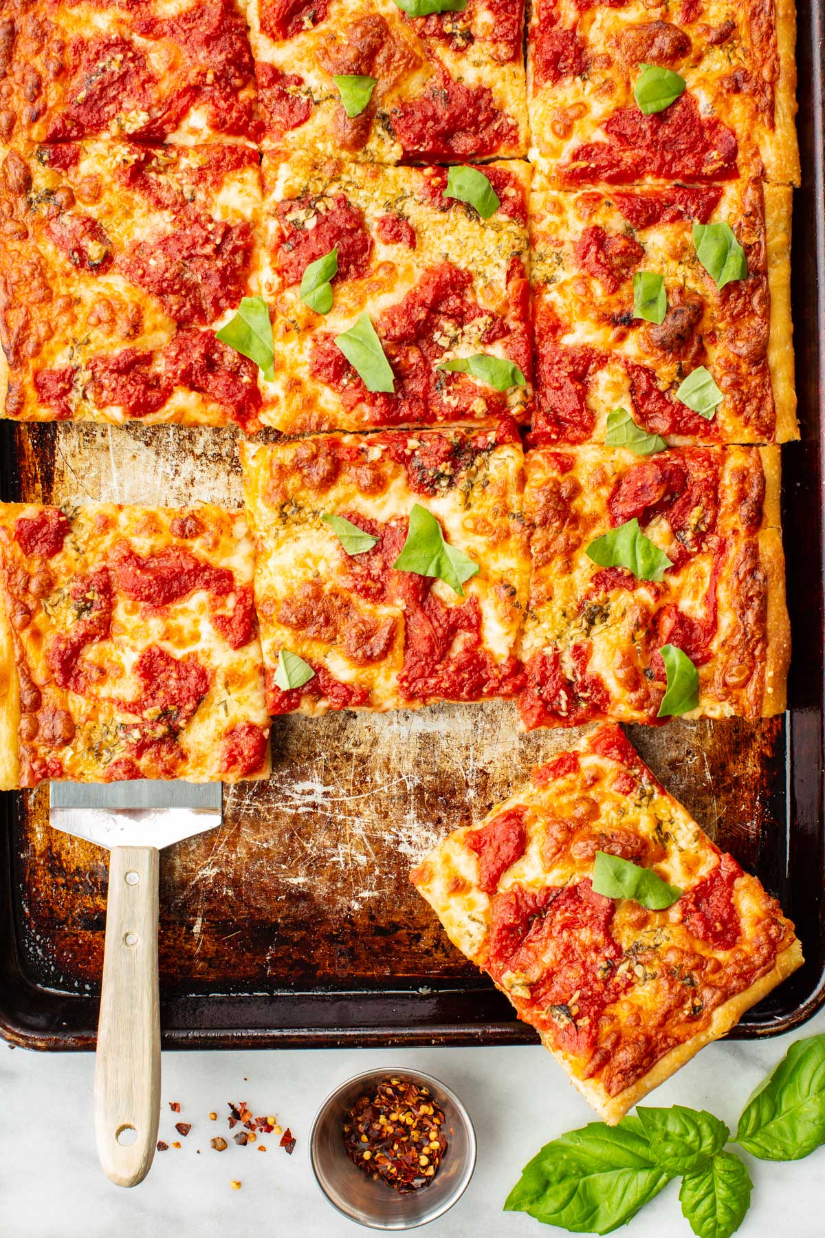 Multiple square slices of grandma pizza arranged on a baking tray and plate, ready to serve, with fresh basil and red pepper flakes alongside.