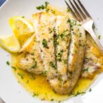 Pan seared white fish fillets spooned with lemon butter sauce, finished with chopped herbs and cracked black pepper, served on a white plate with lemon wedges.