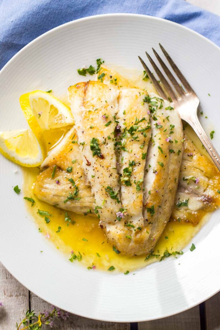 Pan seared white fish fillets spooned with lemon butter sauce, finished with chopped herbs and cracked black pepper, served on a white plate with lemon wedges.