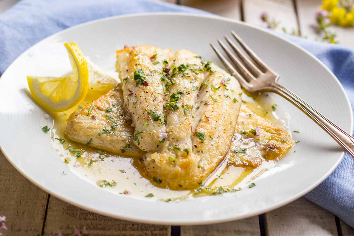 Golden pan seared fish fillets resting in a pool of lemon butter sauce, garnished with fresh herbs and plated with a fork and lemon wedge.
