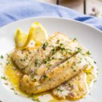 Close up of flaky white fish fillets with crisp golden edges, topped with herbs and bathed in glossy lemon butter sauce.