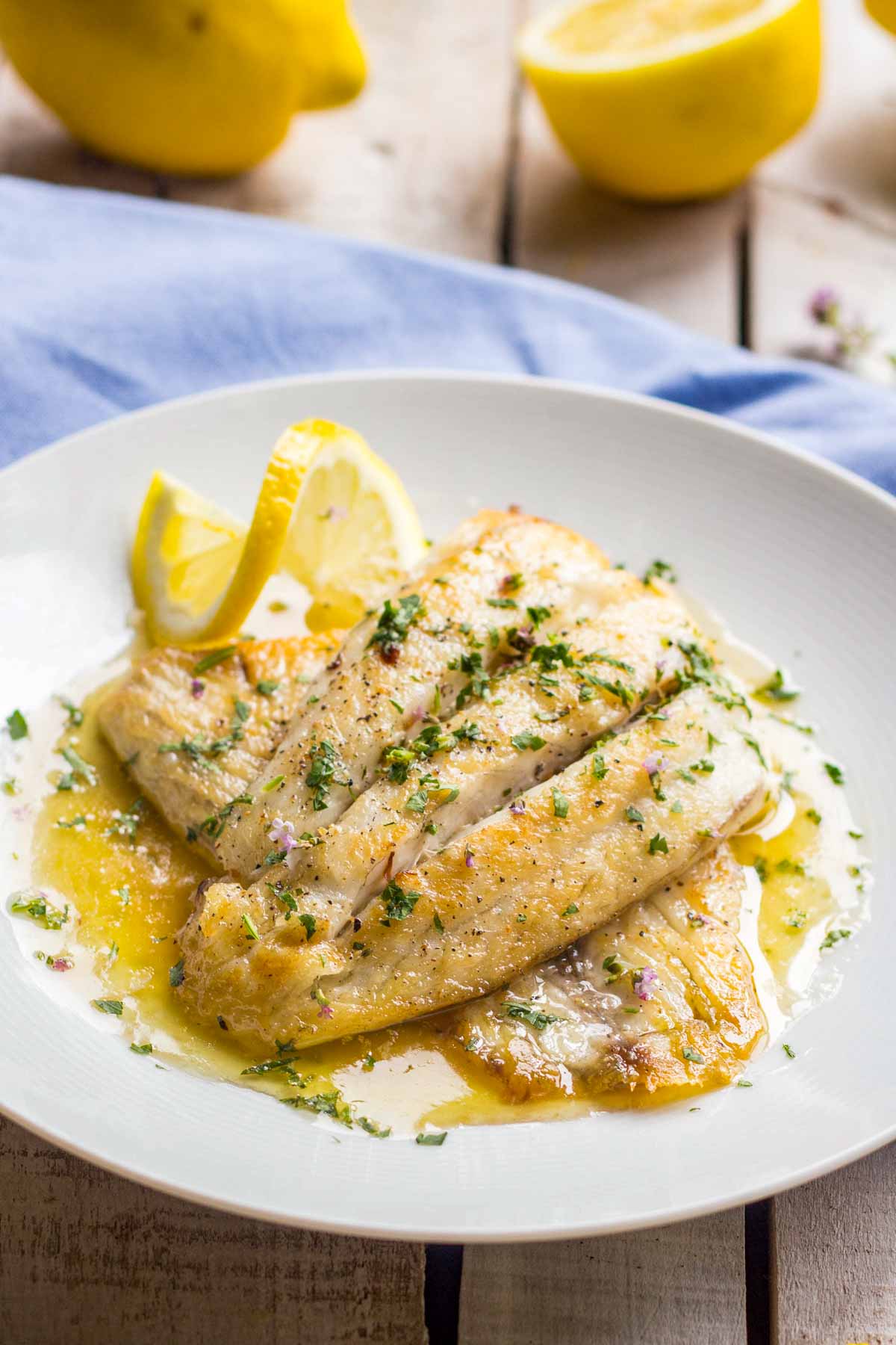 Close up of flaky white fish fillets with crisp golden edges, topped with herbs and bathed in glossy lemon butter sauce.