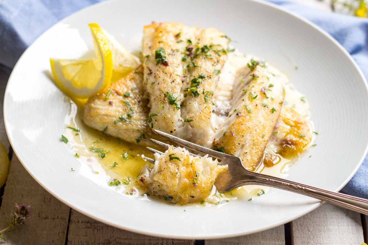 Pan seared fish fillets with a fork pulling away a tender bite, showing the moist flaky interior and lemon butter sauce on the plate.