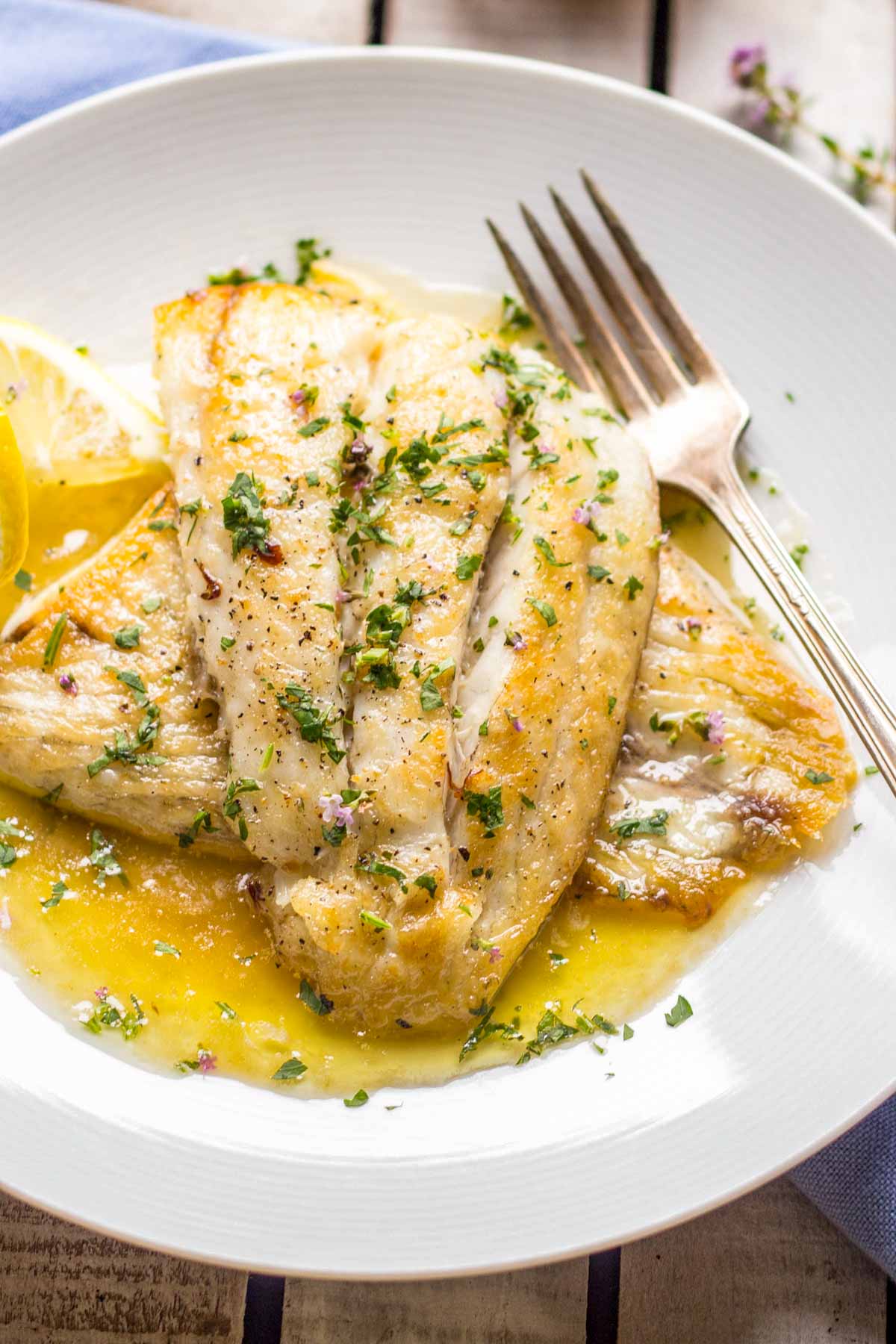 Finished lemon butter fish served on a white plate with fresh herbs, lemon wedges and sauce spooned around the fillets.