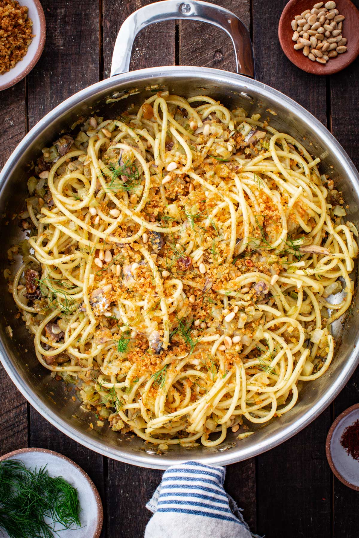Overhead view of pasta con le sarde in a large skillet, spaghetti tossed with sardines, fennel, raisins, pine nuts and toasted breadcrumbs on a rustic wooden table.