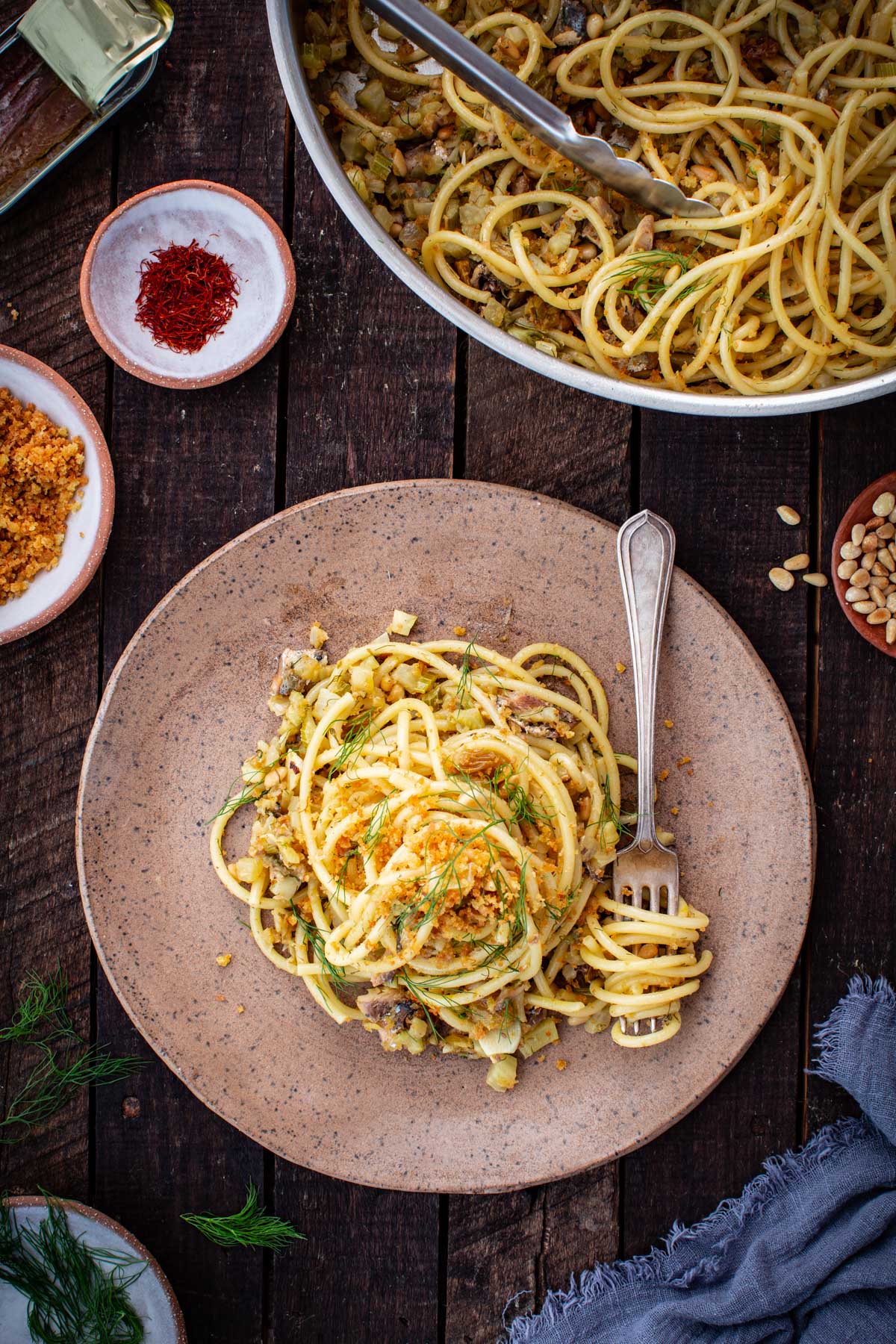 Plated pasta con le sarde topped with golden breadcrumbs and fresh fennel fronds, with the skillet in the background on a dark wood surface.