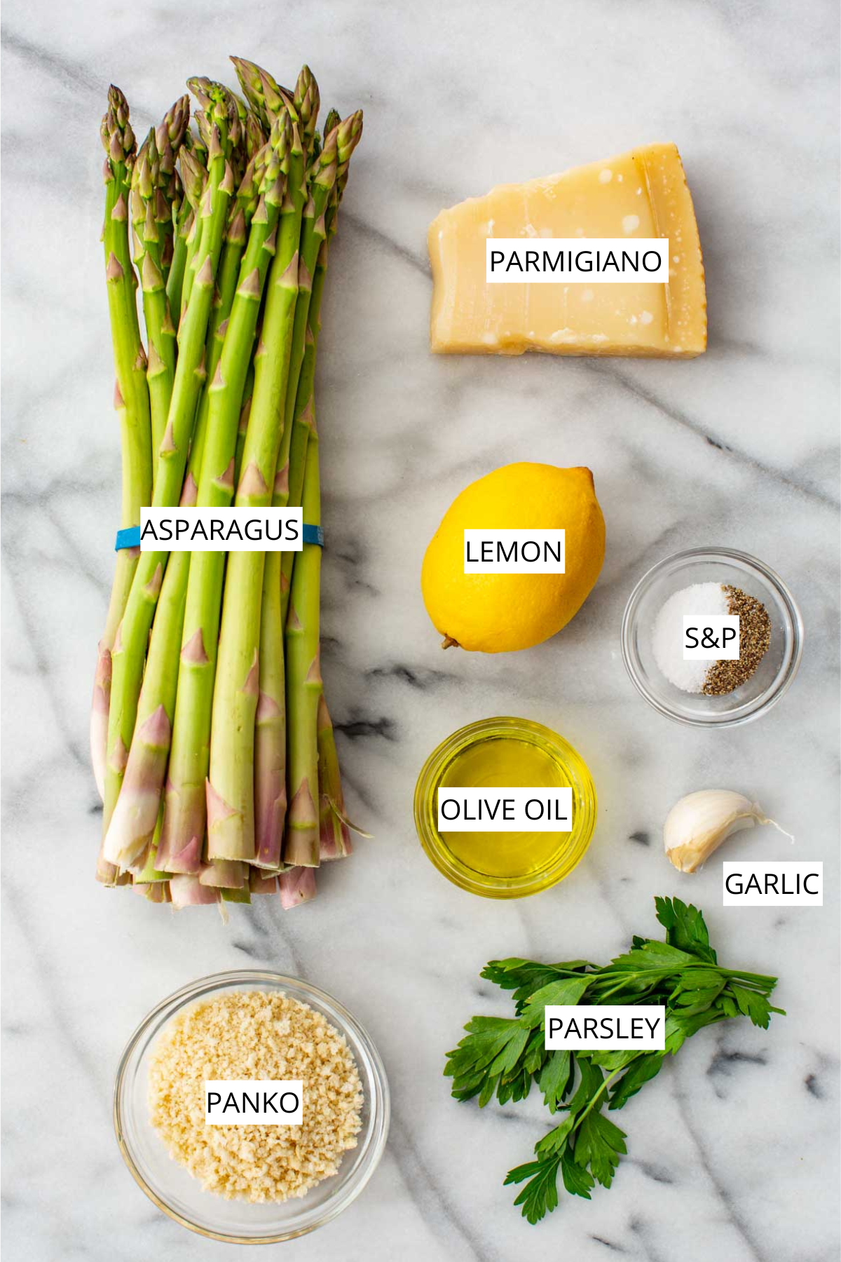 Overhead flat lay of labeled ingredients for parmesan roasted asparagus on a marble surface: a bundle of fresh asparagus, a wedge of Parmigiano, a whole lemon, a small bowl of salt and pepper, a glass jar of olive oil, a garlic clove, a bunch of fresh parsley, and a bowl of panko breadcrumbs.