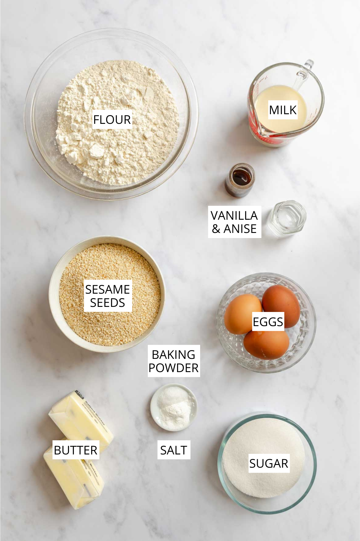 Flat-lay of ingredients for Italian sesame seed cookies on a marble surface, labeled: flour in a large glass bowl, milk in a measuring cup, vanilla and anise extracts in small bottles, sesame seeds in a white bowl, eggs in a glass dish, baking powder in a small dish, butter, salt, and sugar in a glass bowl.
