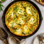 Overhead view of an asparagus ricotta frittata baked in a cast iron skillet, topped with chopped chives and served on a marble surface with small bowls of chives and pepper nearby.