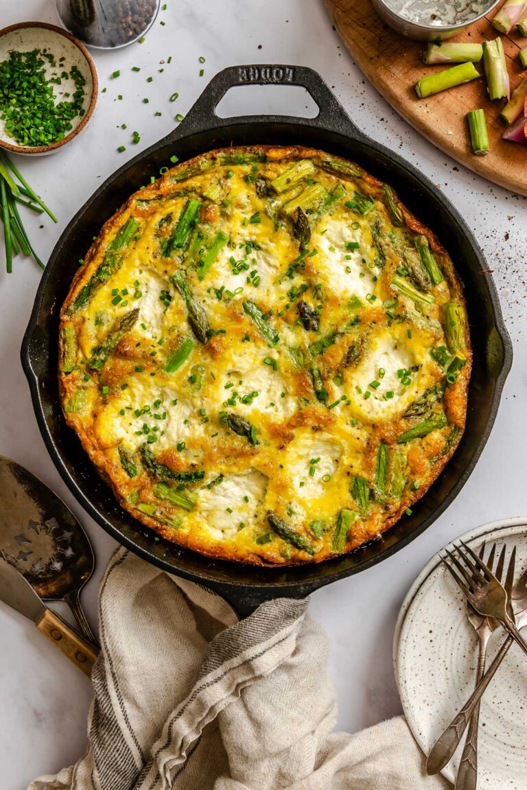 Overhead view of an asparagus ricotta frittata baked in a cast iron skillet, topped with chopped chives and served on a marble surface with small bowls of chives and pepper nearby.