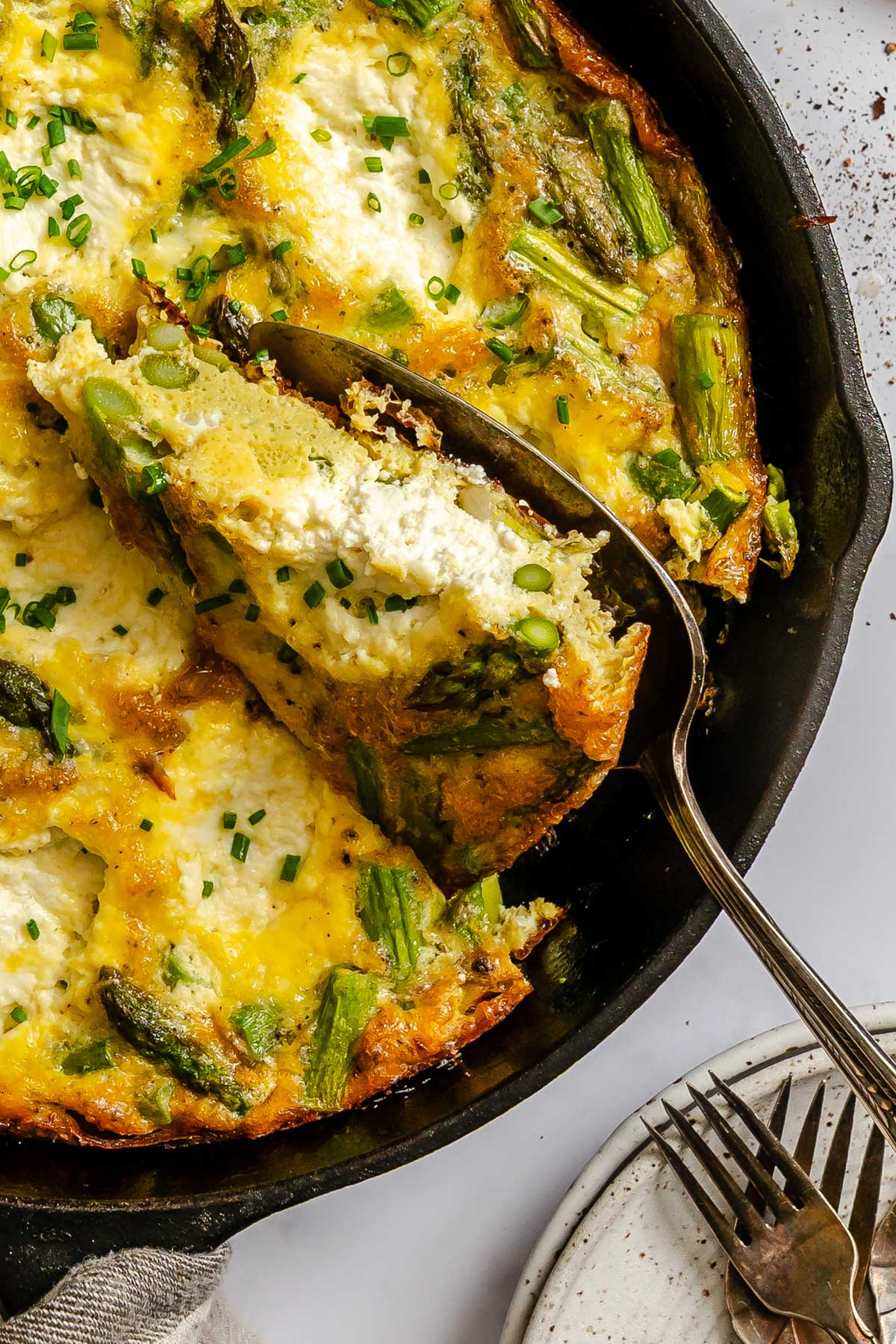 A slice of asparagus ricotta frittata being lifted from a cast iron skillet with a serving spoon, showing fluffy eggs, asparagus and pockets of creamy ricotta.