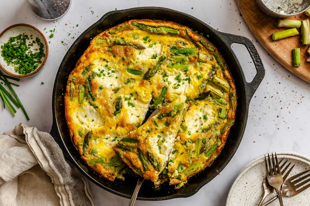 Overhead view of a cast iron skillet with asparagus ricotta frittata as a slice is lifted out, revealing the fluffy egg interior with asparagus and ricotta.