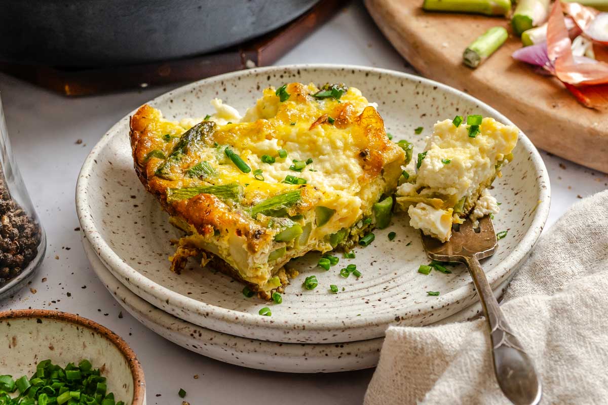 A slice of asparagus ricotta frittata served on a speckled plate with a fork, showing tender asparagus, fluffy eggs and creamy ricotta topped with chopped chives.