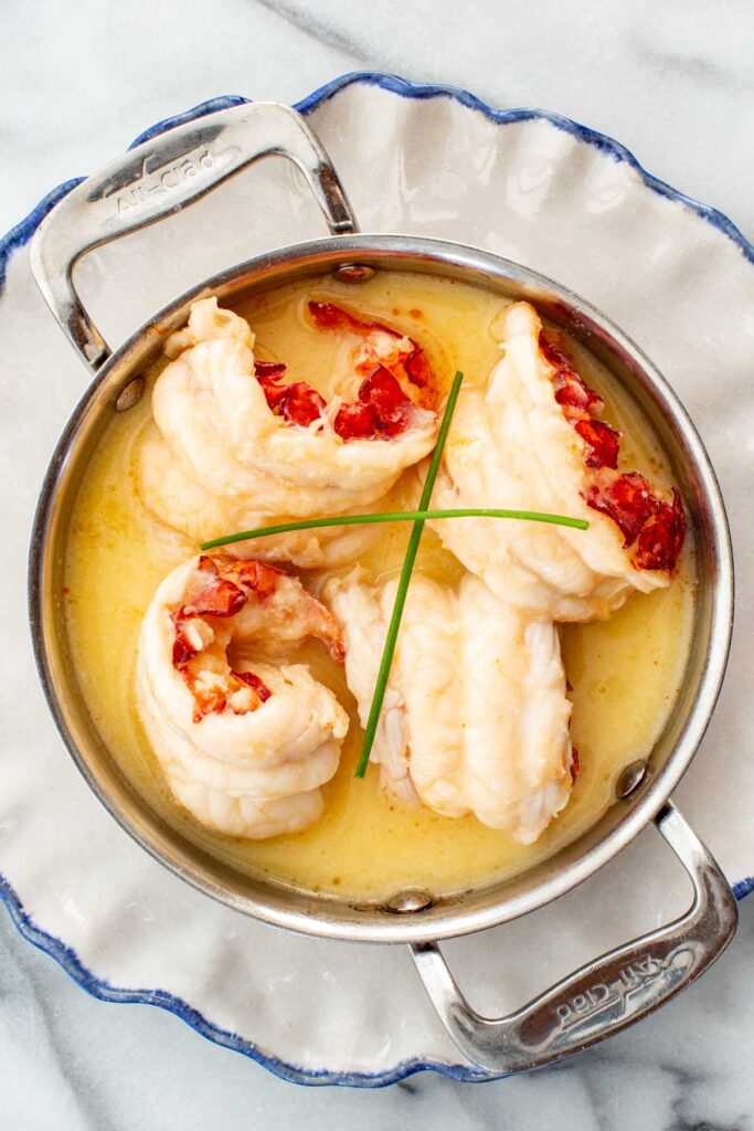 Butter poached lobster plated in a metal pot with two chives sprigs on top.