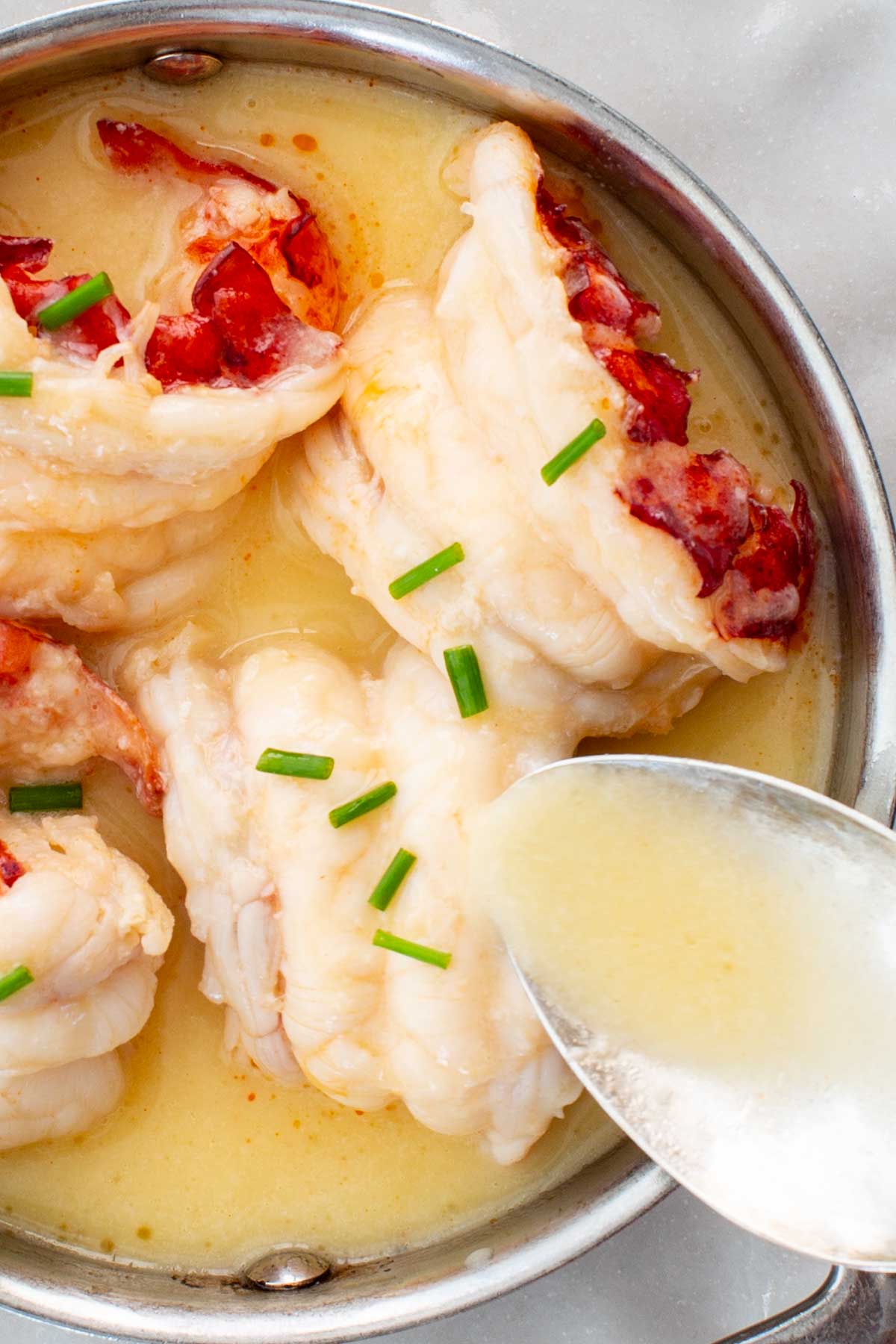 Top view of butter poached lobster in a silver dish with sauce being spooned over lobster tails.