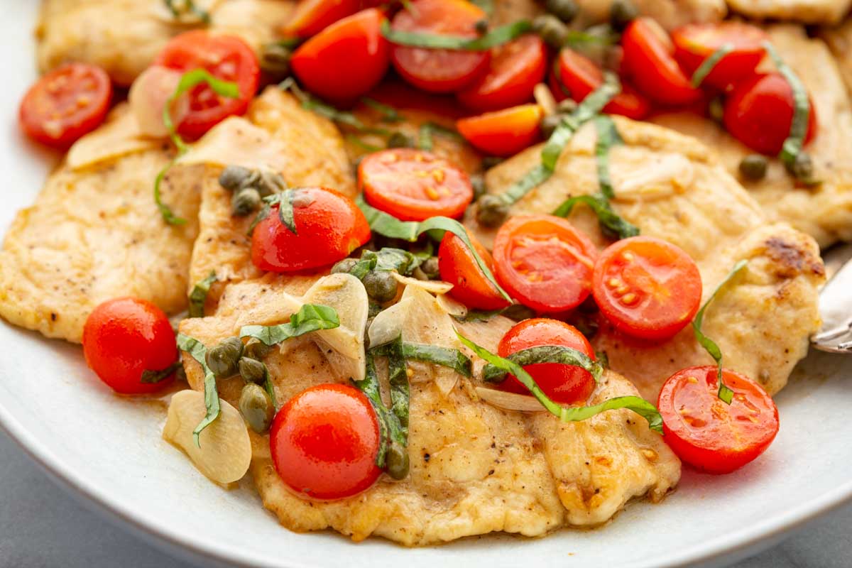 Close-up of golden-brown chicken scallopini on a white plate, topped with halved cherry tomatoes, capers, sliced garlic, and ribbons of fresh basil in a light butter sauce.