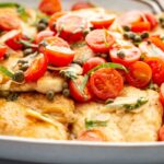 Overhead close-up of chicken scallopini in a large pan, generously topped with halved cherry tomatoes, whole capers, thinly sliced garlic, and fresh basil chiffonade.