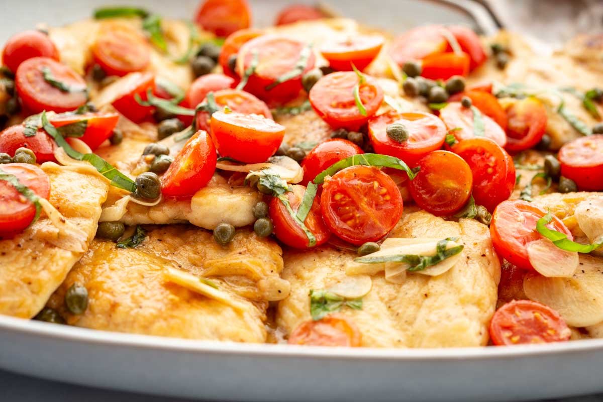 Overhead close-up of chicken scallopini in a large pan, generously topped with halved cherry tomatoes, whole capers, thinly sliced garlic, and fresh basil chiffonade.