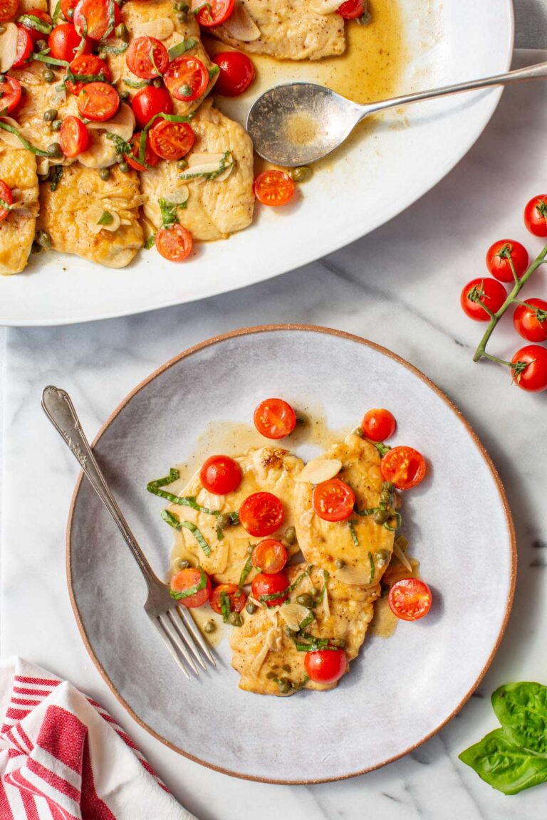 Overhead shot of chicken scallopini served on a large white oval platter with a spoon, alongside an individual portion plated on a round gray dish with a fork, set on a marble surface with fresh cherry tomatoes on the vine and basil leaves.