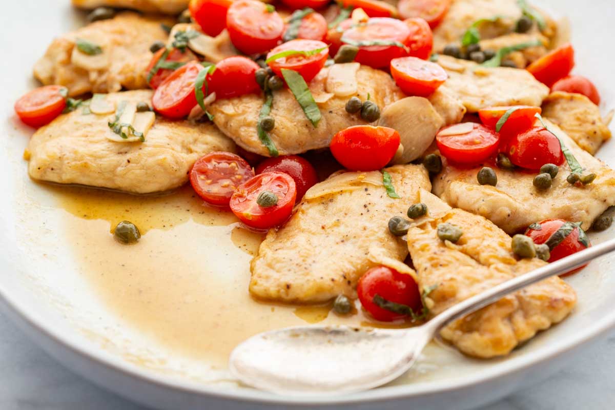 Angled view of chicken scallopini on a white serving platter with a spoon, showing thin pan-seared chicken cutlets in a golden pan sauce topped with halved cherry tomatoes and capers.